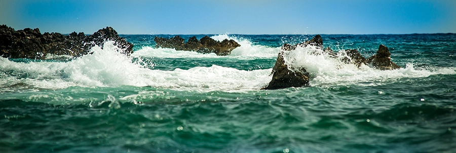 Panoramabild des Meeres, wo kräftige türkisfarbene Wellen mit weißer Gischt an dunkle, zerklüftete Felsen schlagen. Der Himmel ist hellblau, was die Dramatik und Frische der Szene betont. Die Gischt spritzt hoch in die Luft und zeigt die immense Kraft des Ozeans.